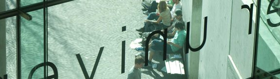 Studierende auf Bänken durch die Frontscheibe der Sowi betrachtet