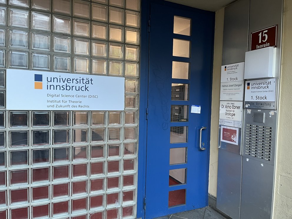 Eingangsbereich mit einer blauen Haustür, links daneben ein großes UIBK-Schild, recht daneben weitere Schilder und Klingeln.