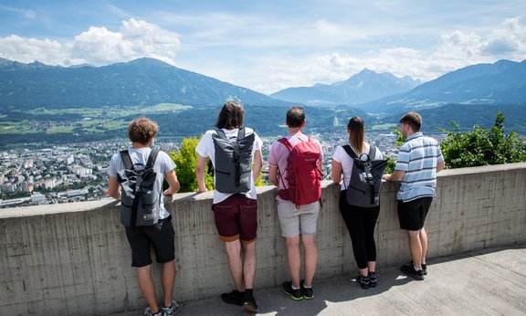 Im Vordergrund das Siegel der Universität Innsbruck, im Hintergrund gehen Menschen in Bewegungsunschärfe durch das Hauptgebäude